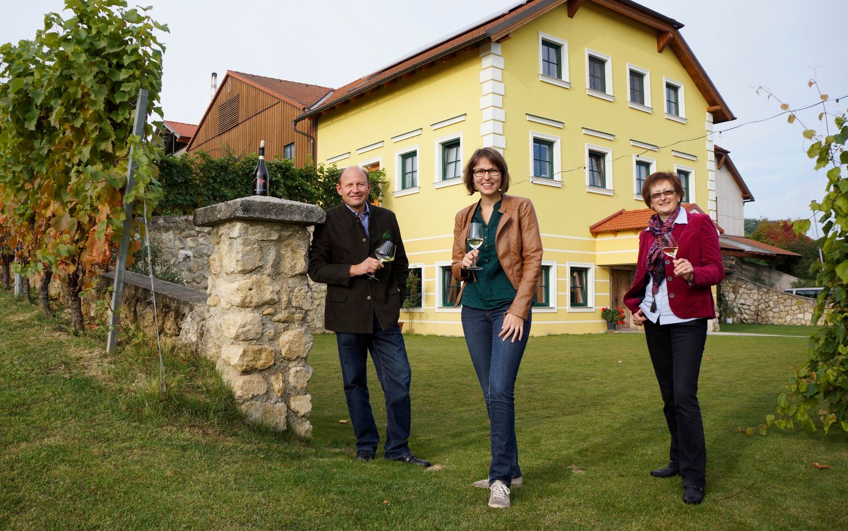 Drei Personen stehen mit Weingläsern vor einem gelben Haus und einem Weinberg.