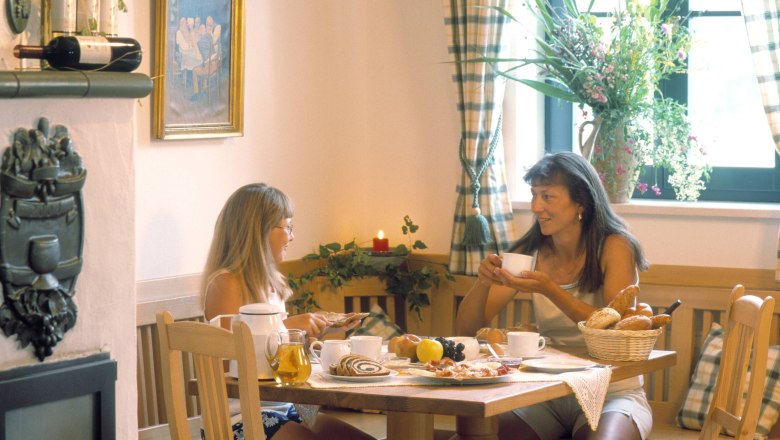 Zwei Frauen frühstücken an einem Tisch in einem gemütlichen Raum mit Blumen und Weinflaschen.