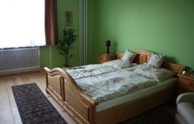 Ein gemütliches Gästezimmer mit grün gestrichenen Wänden, einem Holzbett, Nachttischen, einer Pflanze und einem Teppich auf Holzboden.