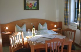 Gemütliches Gästezimmer mit Doppelbett, Tisch und Stühlen, Weinflasche und Gläsern, Fenster mit Vorhängen.