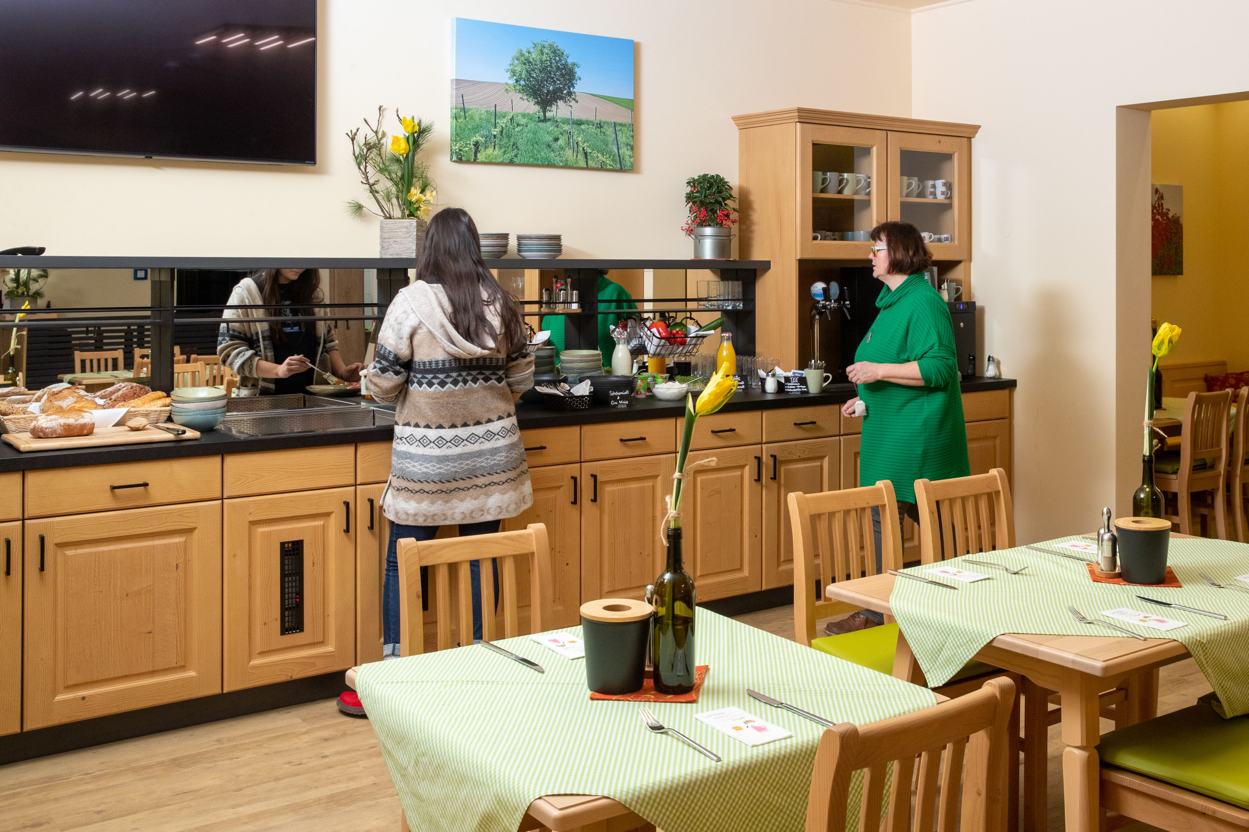 Zwei Personen bedienen sich an einem Frühstücksbuffet in einem gemütlichen Raum mit Holzmöbeln und grünen Tischdecken.