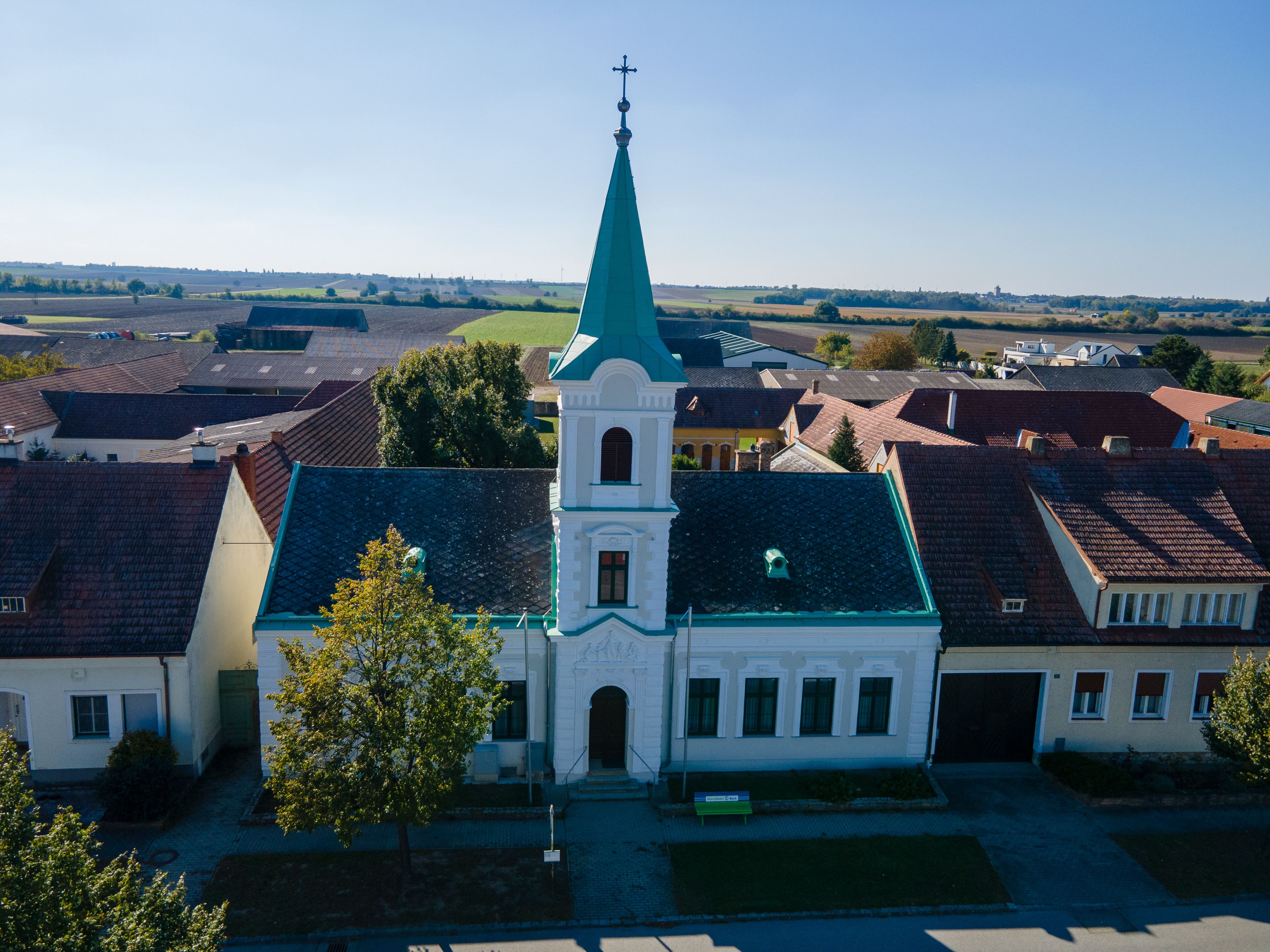 Kirche in der Gemeinde Weiden an der March mit grünem Turm und Kreuz, umgeben von Häusern und Feldern.