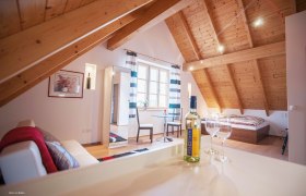 Gemütliches Dachgeschosszimmer mit Holzbalkendecke, Bett, Sofa, Tisch und Weinflasche.