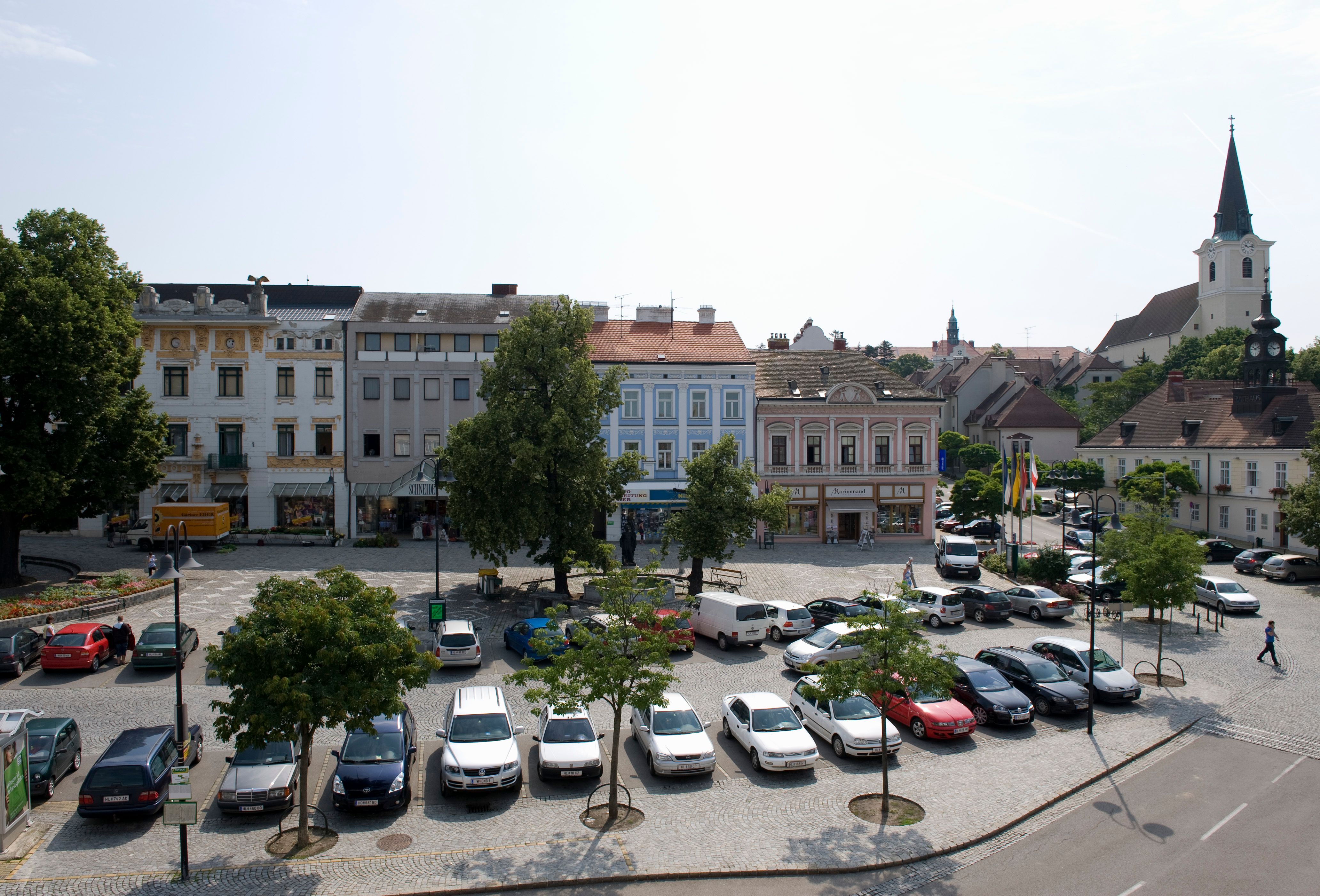 Stadtplatz in Hollabrunn mit geparkten Autos, historischen Gebäuden und einer Kirche im Hintergrund.