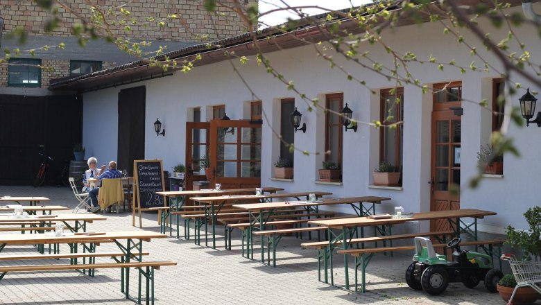 Innenhof einer Buschenschank mit leeren Holzbänken und einem Tisch, zwei Personen sitzen an einem Tisch, ein Spielzeugtraktor steht daneben.