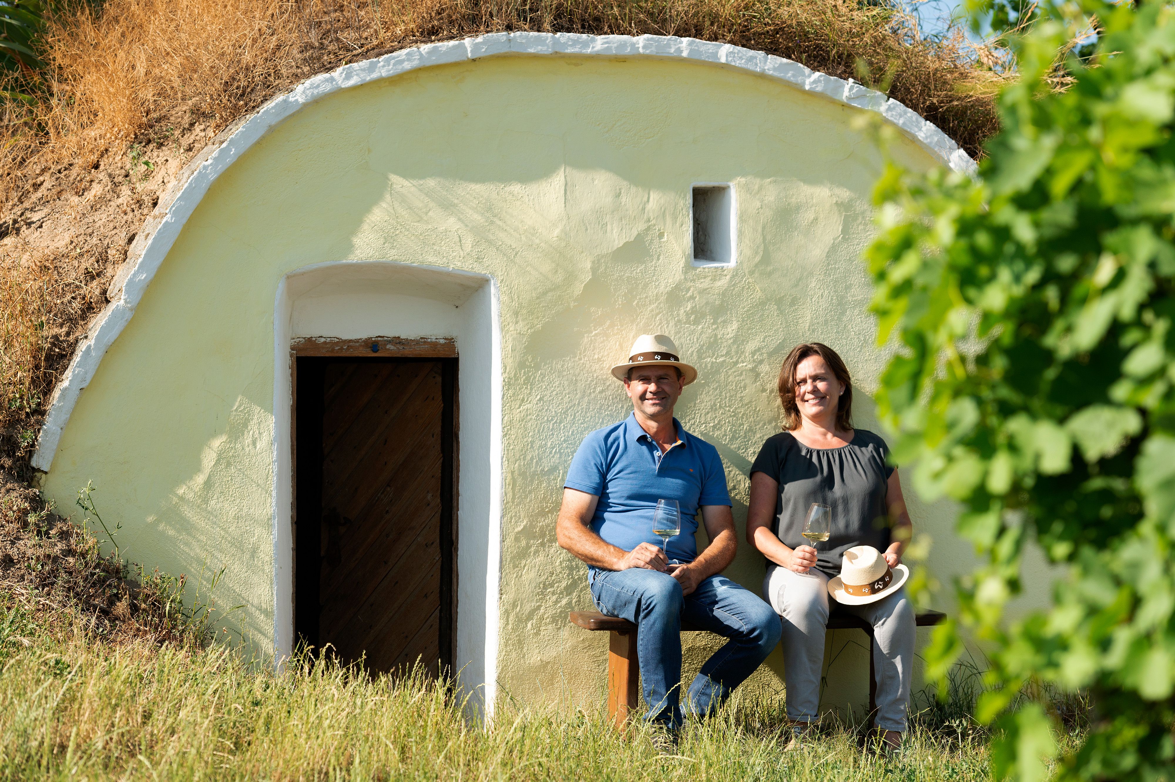 Two people are sitting in front of a hut with glasses of wine in their hands.