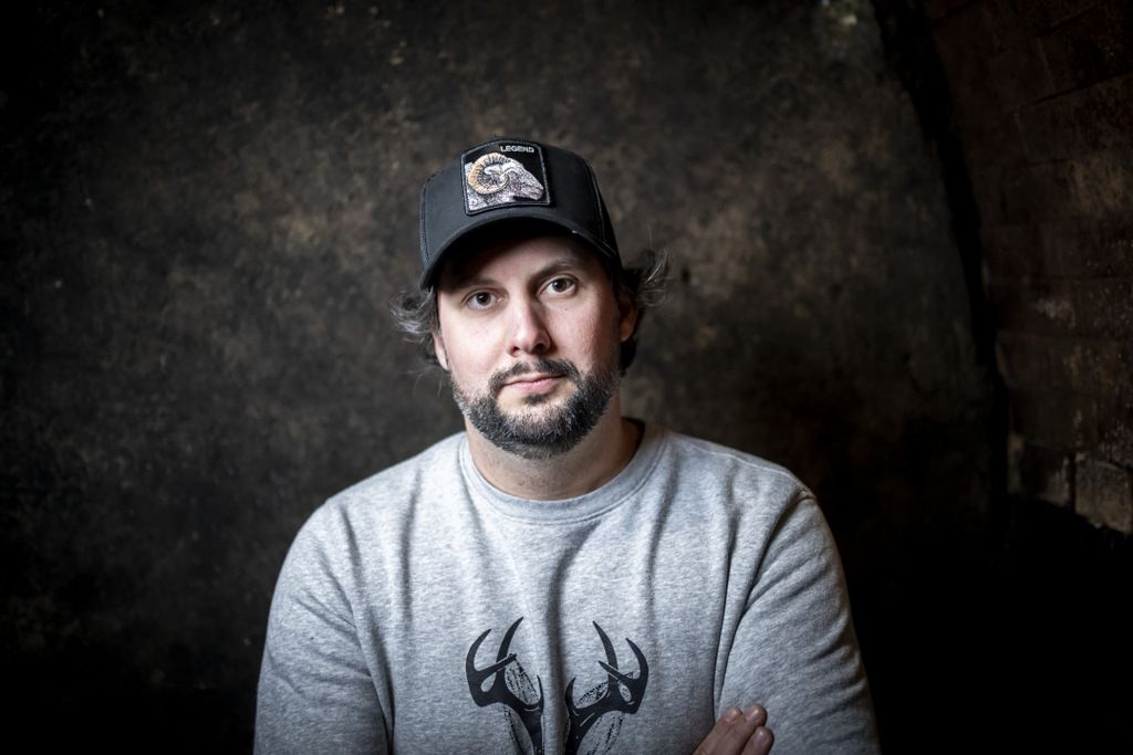 Ein Mann mit Bart und Baseballkappe steht vor einer dunklen Wand.