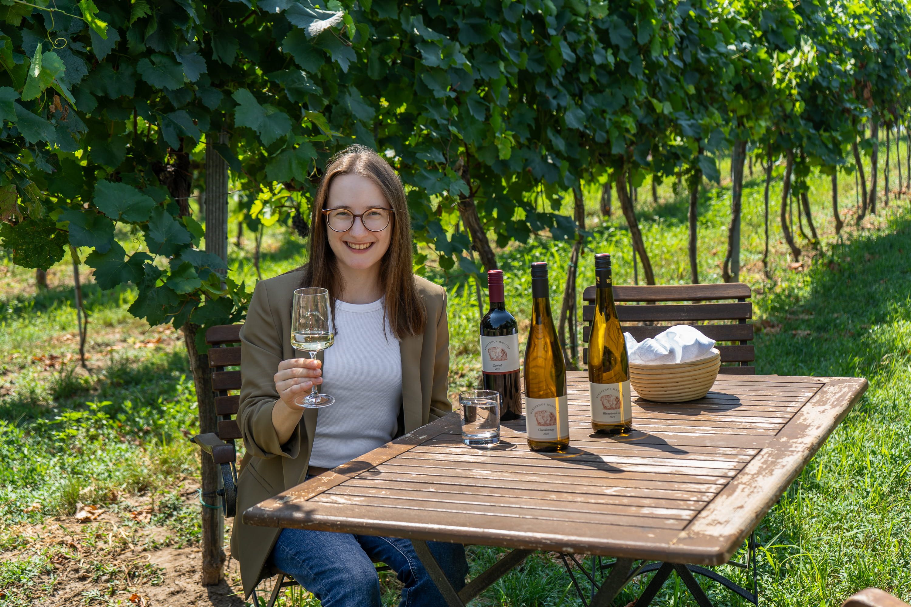A woman is sitting at a wooden table in the vineyard holding a wine glass. There are several bottles of wine on the table.