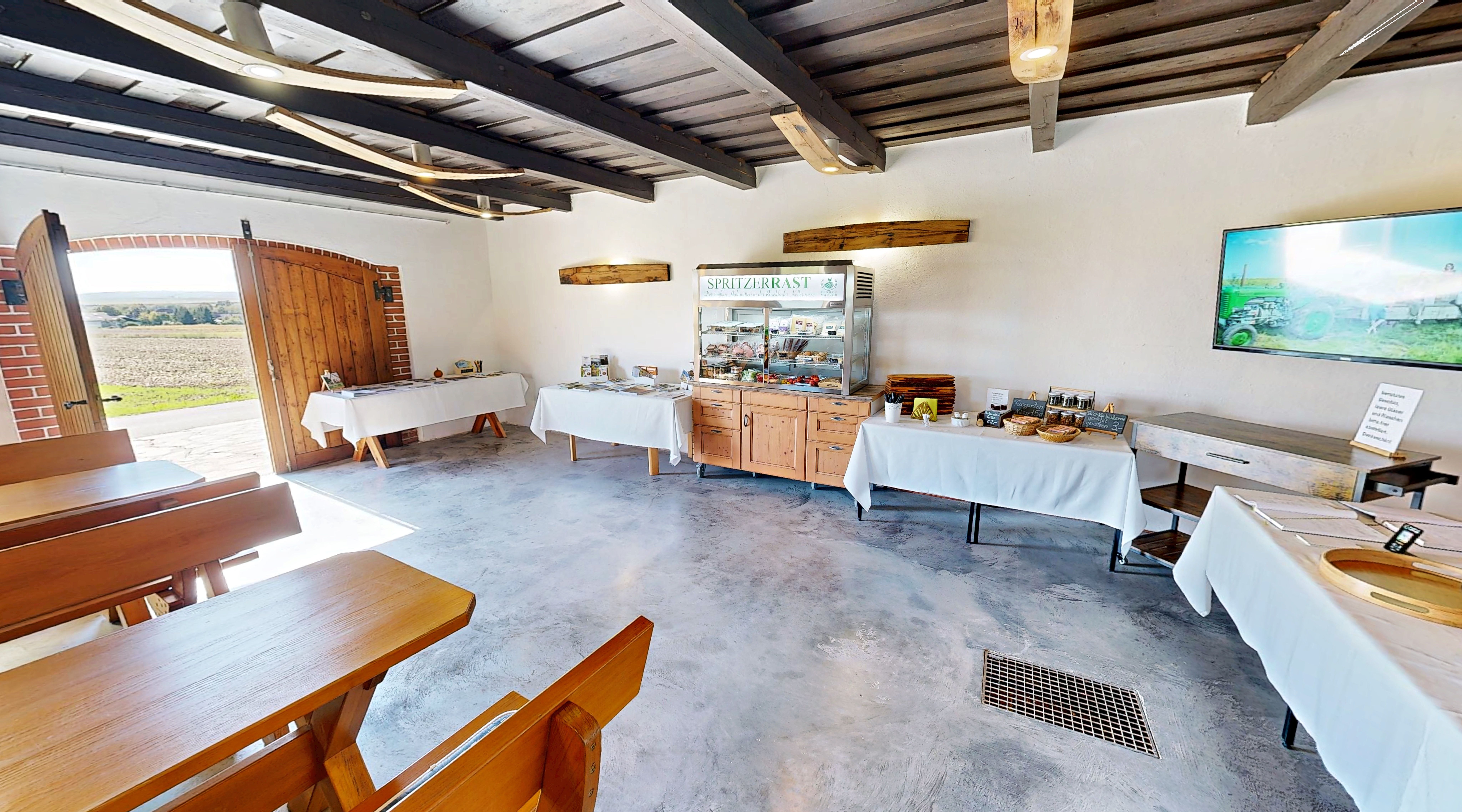 Innenansicht eines Raumes mit Holzbänken, Tischen und einem Buffet mit Speisen. Eine offene Tür bietet Blick auf eine Landschaft.