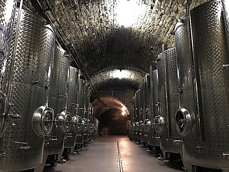 A wine cellar with large stainless steel tanks in a vaulted, stone room.