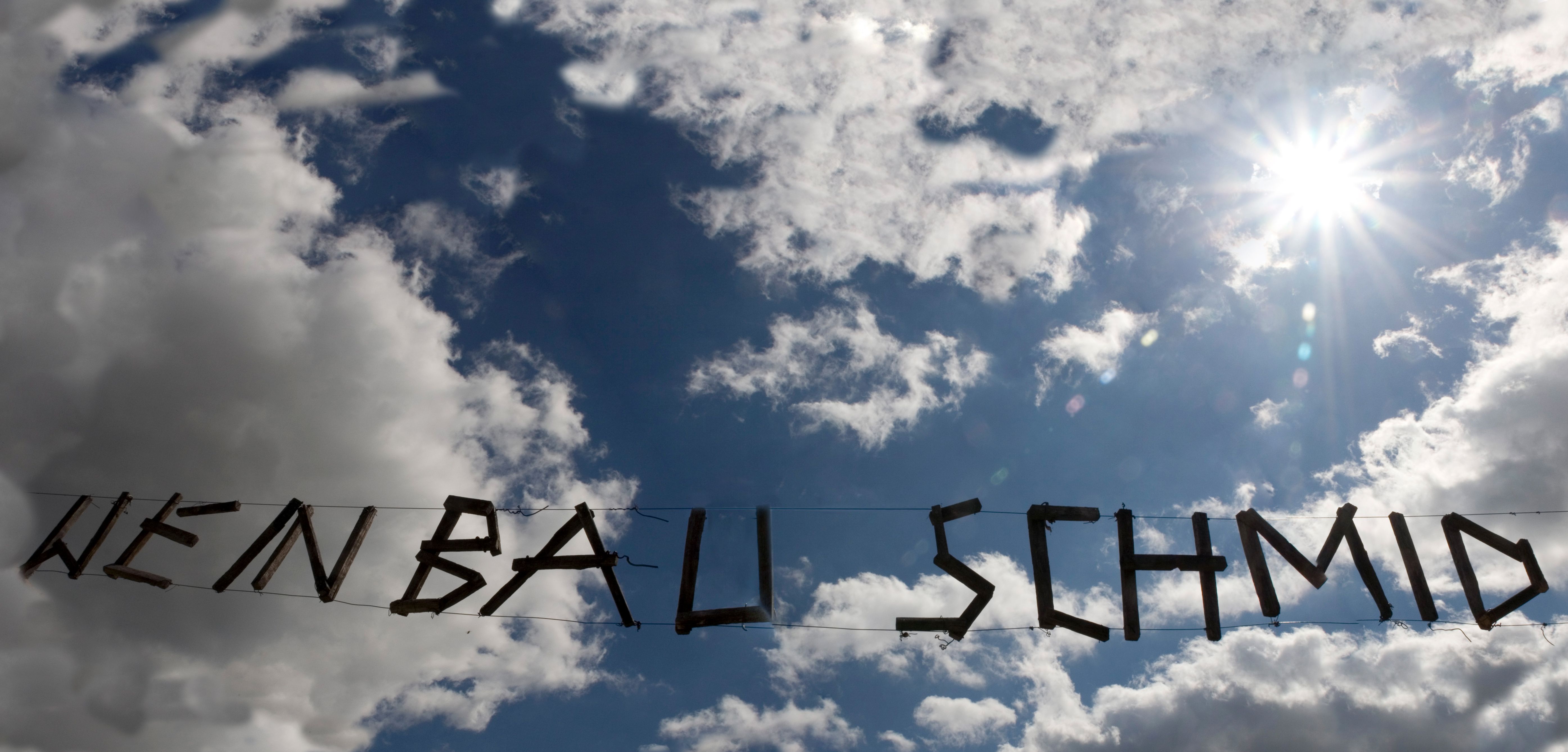 Wooden lettering 'Weinbau Schmid' in front of a cloudy sky and sun.