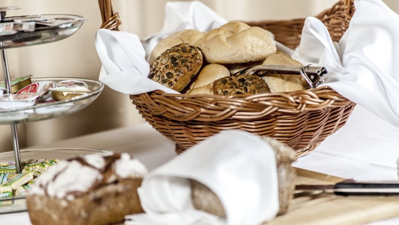 A breakfast table with a basket full of bread rolls, a bread cutting board and a tray of jams.