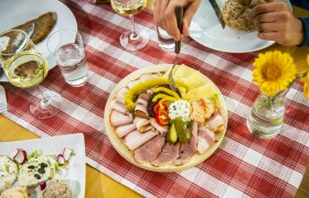 Ein gedeckter Tisch mit einer Platte voller Aufschnitt, Käse und Gemüse, daneben Gläser mit Weißwein und Wasser auf einer rot-weiß karierten Tischdecke.