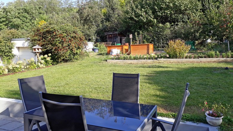 Terrasse mit Tisch und Stühlen, Blick auf einen Garten mit Rasen, Bäumen und Hochbeeten.