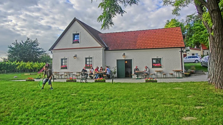 A traditional building with a red roof, surrounded by a green meadow and trees, people sitting at tables outside.