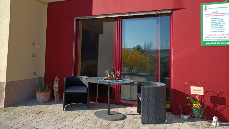 Terrasse mit zwei Stühlen und einem Tisch vor einer roten Wand, daneben ein Schild mit Informationen.