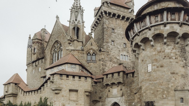 Two people are standing in front of Kreuzenstein Castle, surrounded by old stone walls and towers.