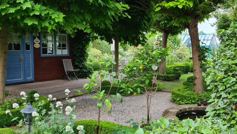 An idyllic garden with a small wooden house, surrounded by trees and plants, with a gravel path and a small pond.