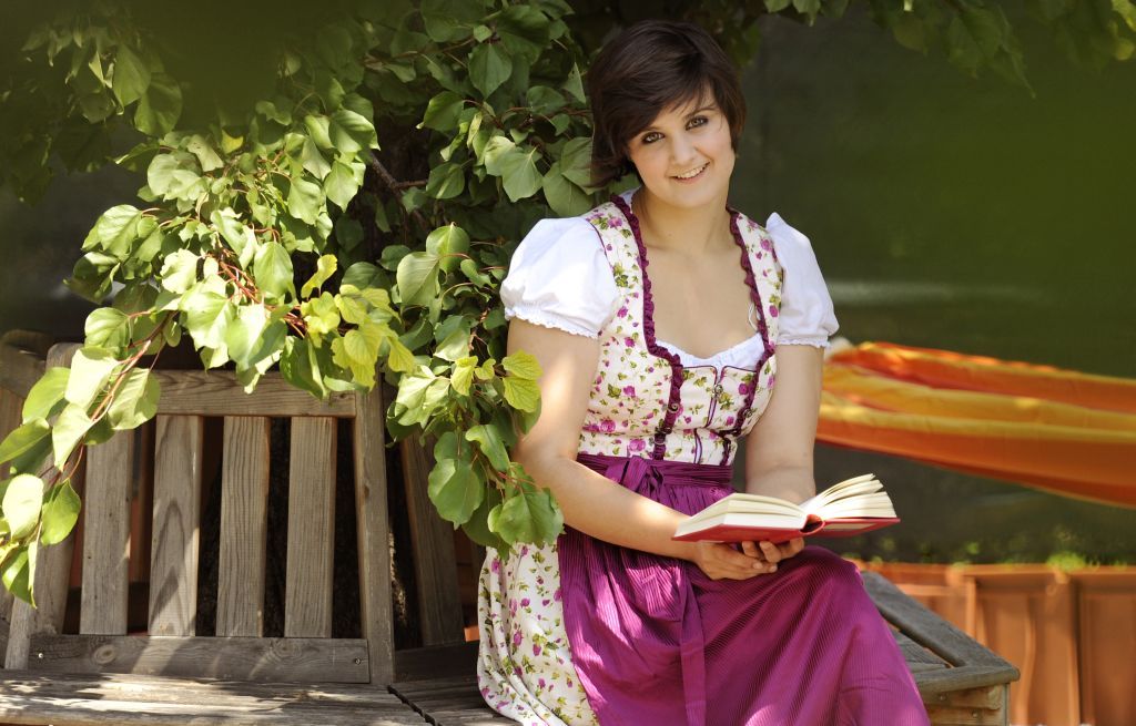 Frau in traditioneller Kleidung liest ein Buch auf einer Holzbank unter einem Baum.