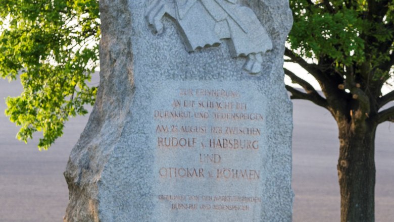 Gedenkstein mit Ritterrelief und Inschrift, umgeben von Bäumen und Wiese.