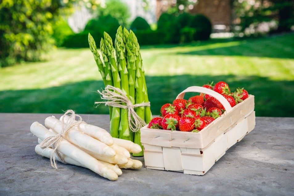 Ein Bund grüner und weißer Spargel neben einem Korb voller Erdbeeren auf einem Tisch im Freien.