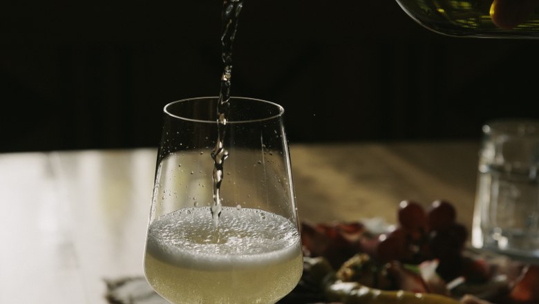 A glass of white wine is poured on a table, with a plate of snacks in the background.