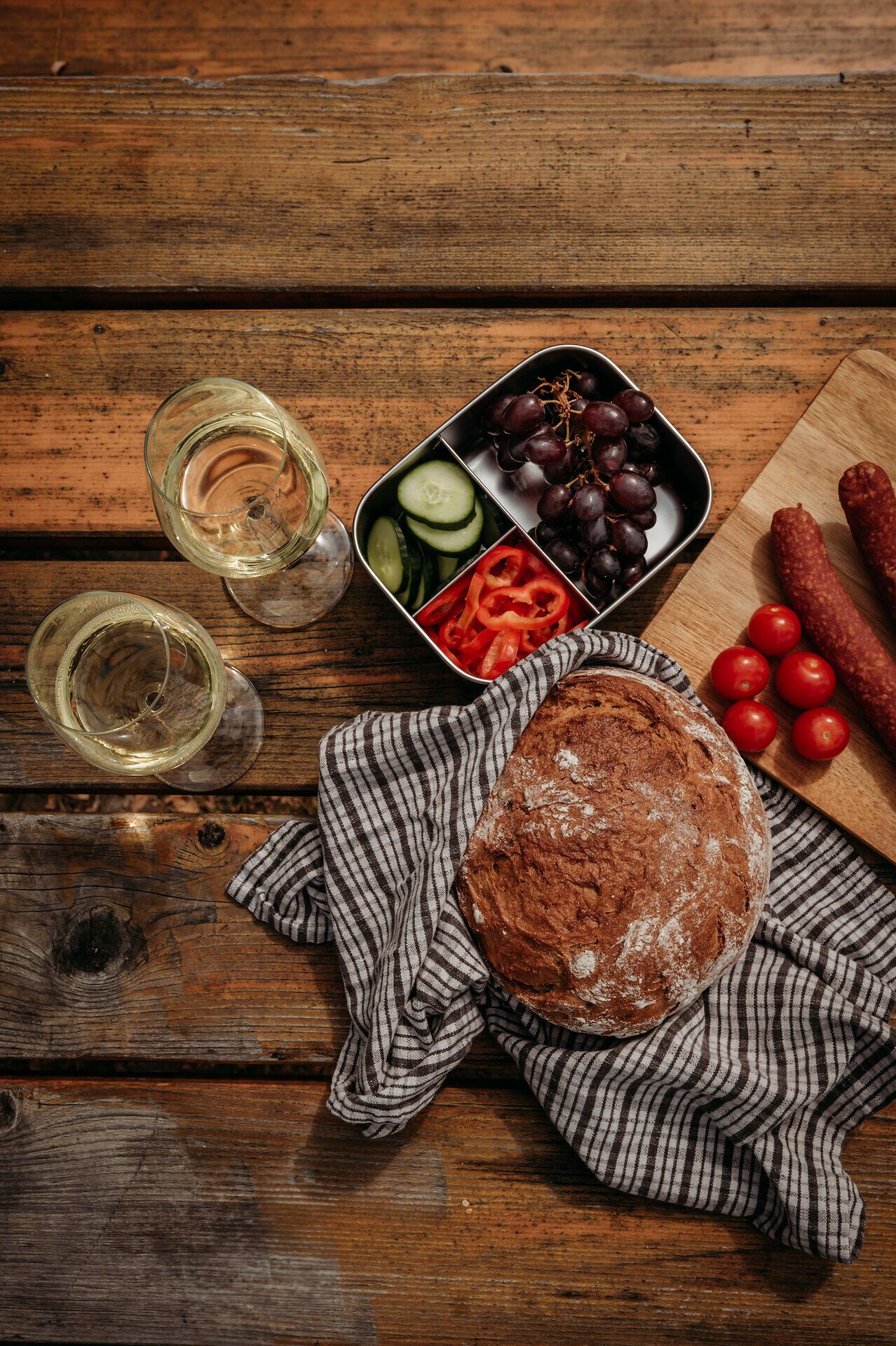 Auf einem Holztisch ist eine Jause mit zwei Gläsern Weißwein, einem Laib Brot, einer Box mit Trauben und Gemüse sowie einem Holzbrett mit Weinviertler Hartwurst angerichtet.