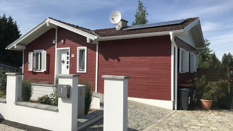 Red wooden house with white shutters and solar panels on the roof, surrounded by a paved courtyard and a low wall.
