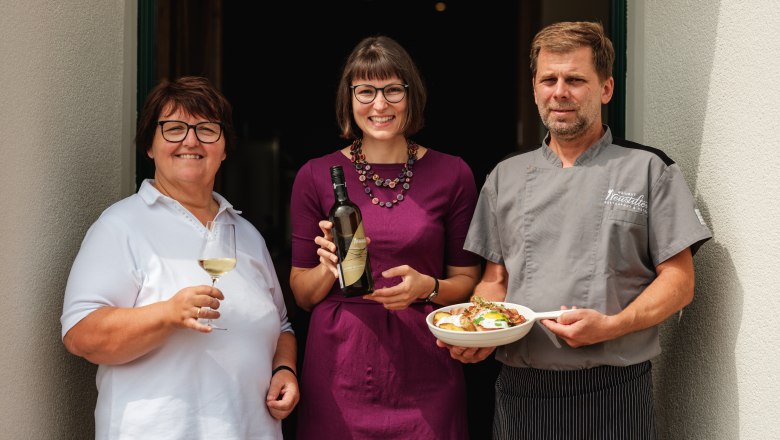 Three people in front of a restaurant, a woman with a wine glass, a woman with a wine bottle, a man in a chef's outfit with a plate.