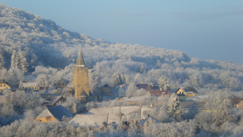 Winterlandschaft mit Dorf und Kirche, umgeben von schneebedeckten Bäumen.