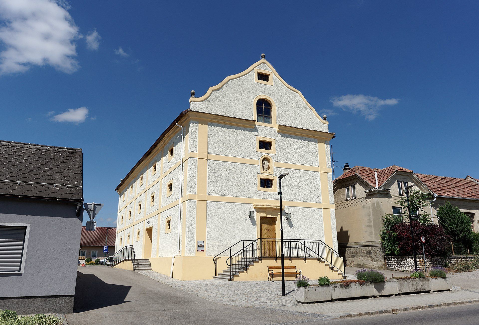 Historická budova so žltými akcentmi a schodiskom, obklopená ďalšími domami pod modrou oblohou.