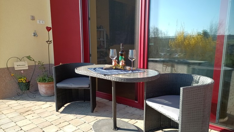 Terrasse mit Tisch, zwei Stühlen, Weinflasche und Gläsern vor einer roten Tür.