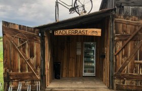Holzhütte mit Schild 'Radlerrast', Fahrrad auf dem Dach, Getränke im Inneren.