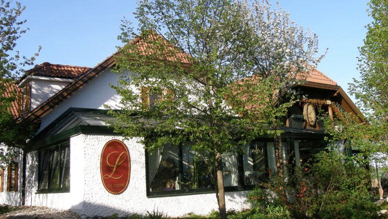 Außenansicht eines traditionellen Gebäudes mit roten Dachziegeln und einem großen L-Logo an der Wand, umgeben von Bäumen und Pflanzen.