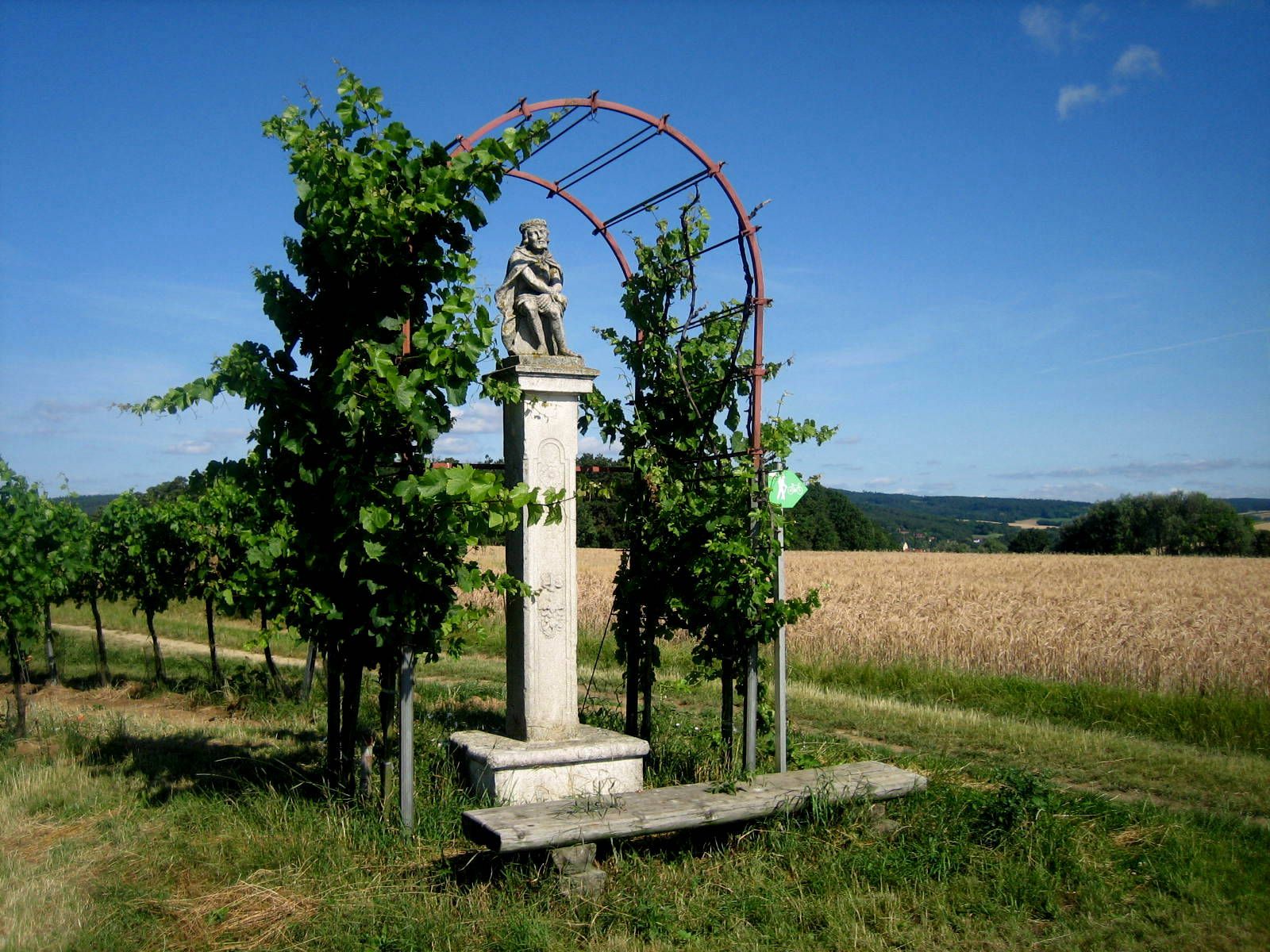 Steinernes Kreuz mit Figur in ländlicher Umgebung, umgeben von Pflanzen und Feldern.
