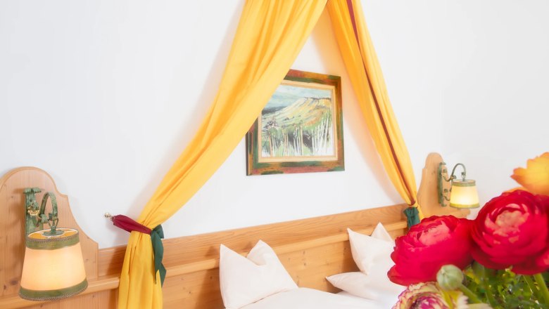 Ein gemütliches Doppelzimmer mit gelbem Baldachin, Holzbett, Wandlampen und einem Blumenstrauß im Vordergrund.