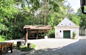 Ein gepflasterter Hof mit einem kleinen weißen Gebäude und einem überdachten Holzgestell, umgeben von Bäumen.