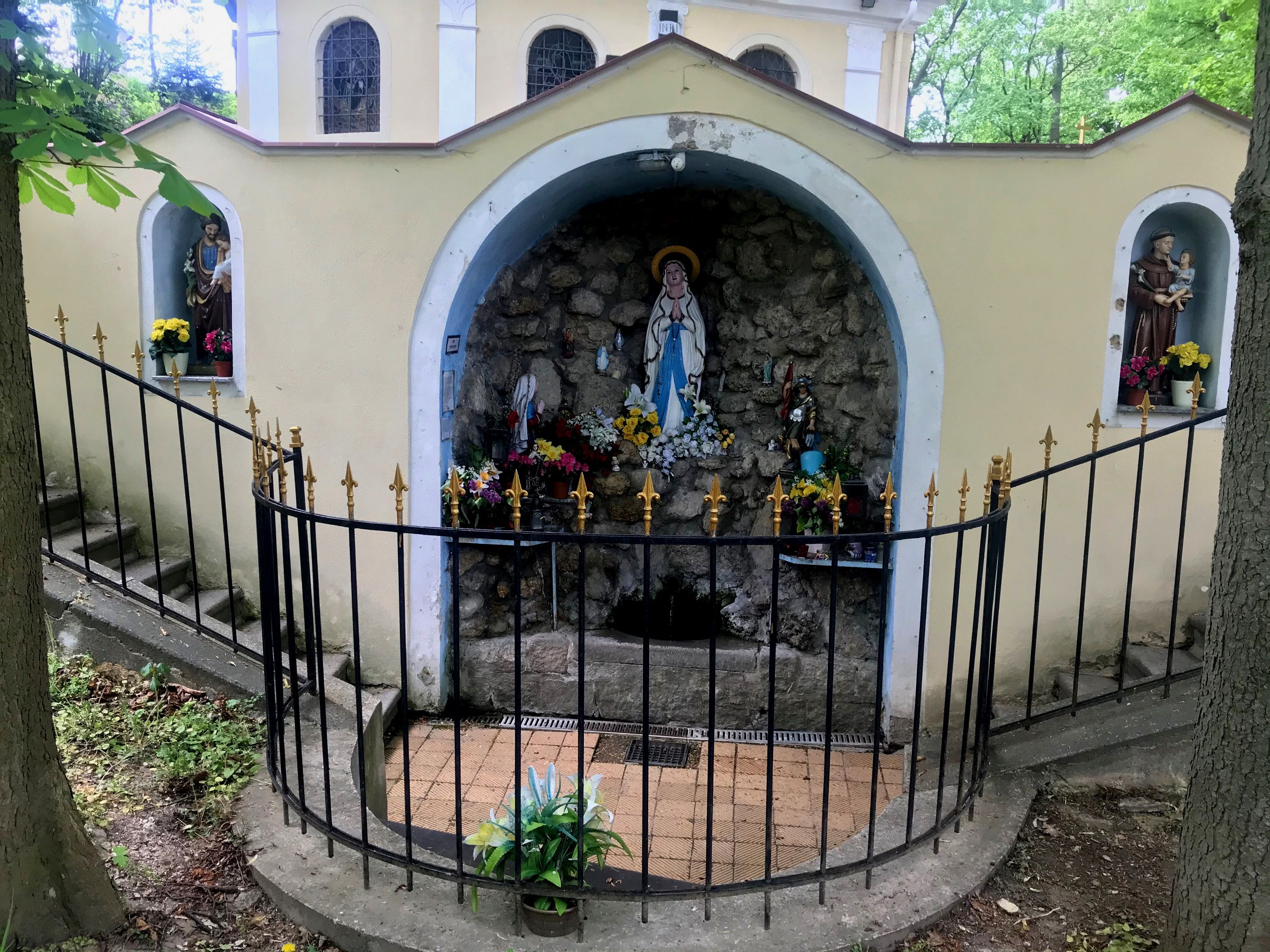 Grotta s náboženskými sochami a kvetmi, obklopená kovaným plotom.