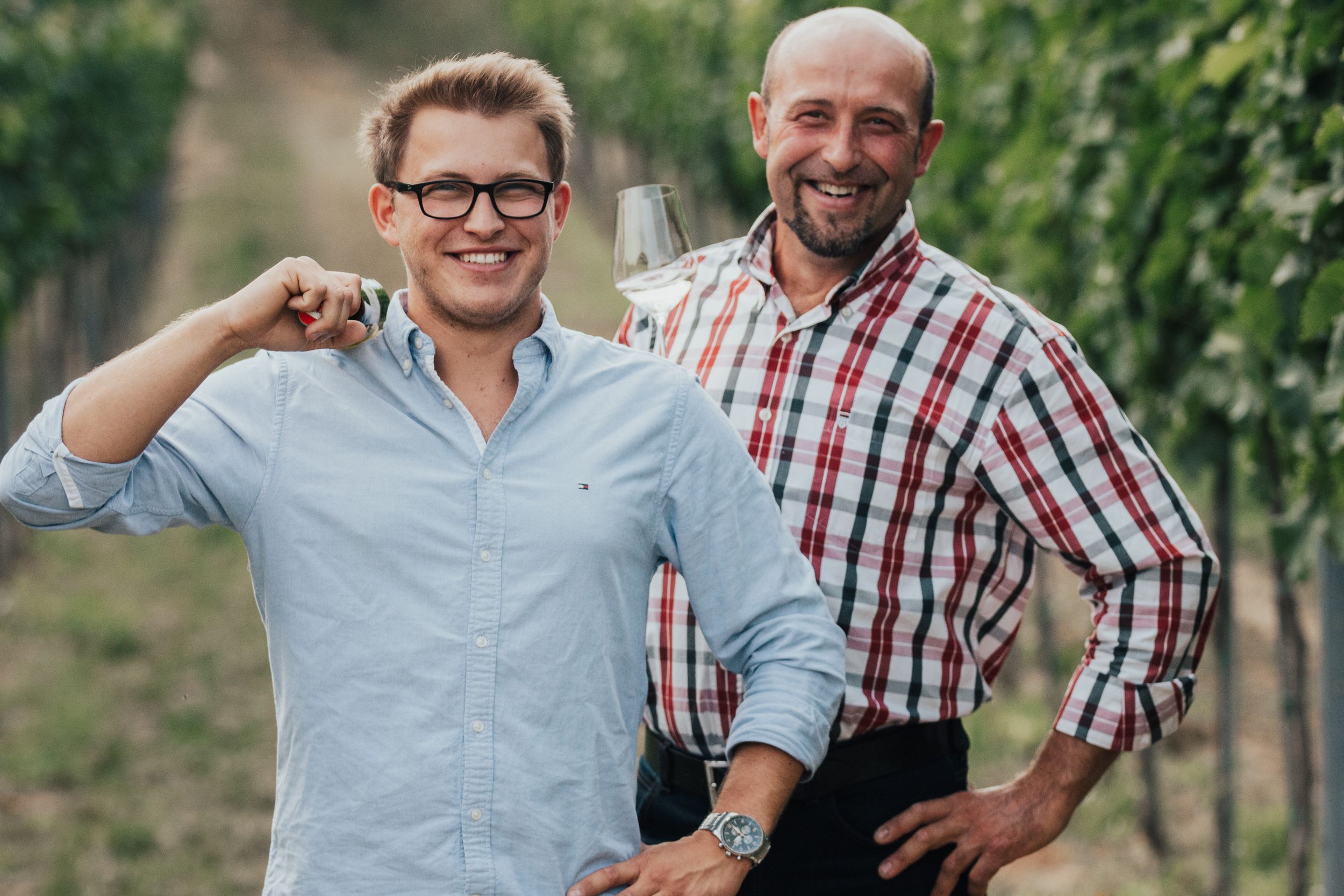 Two men stand smiling in a vineyard, one holding a wine glass.