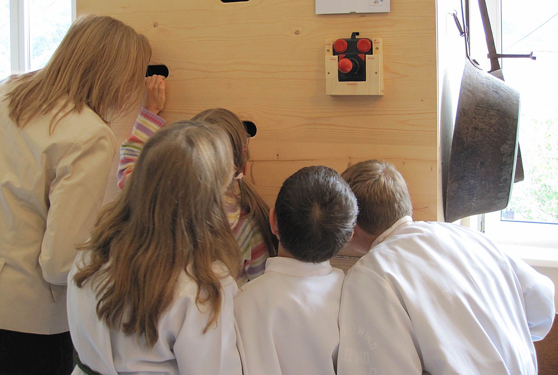 Group of children and a woman looking through holes in a wooden wall.