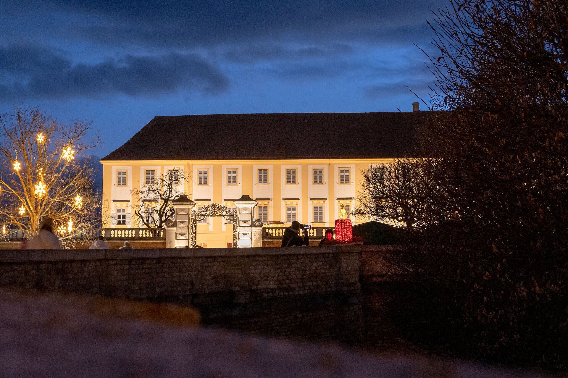 In der Dämmerung erstrahlt das Schloss Hof in warmem Licht und schafft eine festliche Atmosphäre. Die umstehenden Bäume sind mit funkelnden Lichtern geschmückt, während Besucher den Adventmarkt erkunden und die Vorfreude auf Weihnachten spüren. Ein Ort, der die Herzen der Gäste mit seiner zauberhaften Kulisse verzaubert.