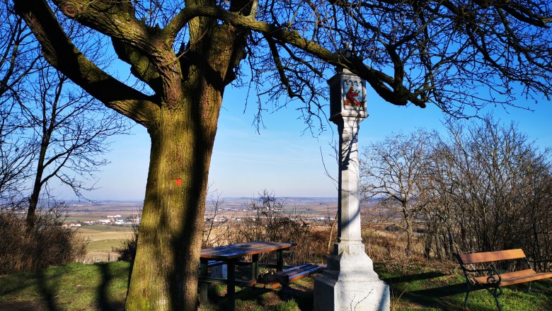 Ein gro&szlig;er Baum neben einem steinernen Wegkreuz mit B&auml;nken und einem weiten Ausblick auf die Landschaft.