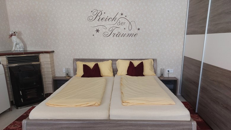 Ein Schlafzimmer mit einem Doppelbett, gelber Bettwäsche und einem Schriftzug an der Wand. Ein Ofen und eine Hundeskulptur sind ebenfalls zu sehen.