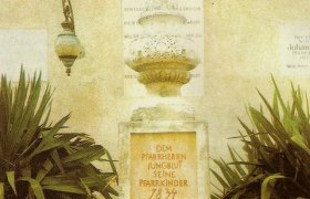 Denkmal mit Inschrift, umgeben von Pflanzen und einem Korb mit Kartoffeln und Sonnenblumen.