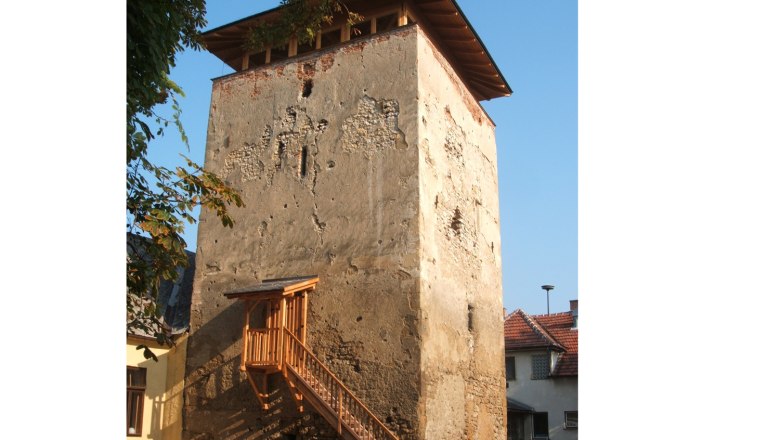 Ein alter Wehrturm mit bröckelnder Fassade und einem hölzernen Treppenaufgang, umgeben von Häusern und Bäumen.