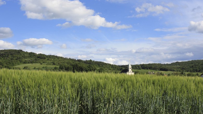 Landschaft mit grünem Feld, Kirche im Hintergrund und blauem Himmel mit Wolken.
