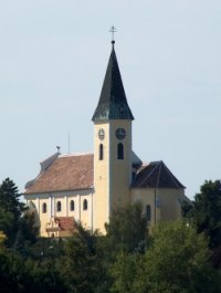 Pfarrkirche Großebersdorf mit spitzem Turm und Uhr, umgeben von Bäumen.