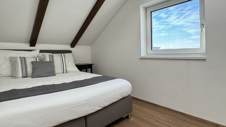 Modernes Schlafzimmer mit Holzbalkendecke, großem Bett und Fenster mit Blick auf blauen Himmel.