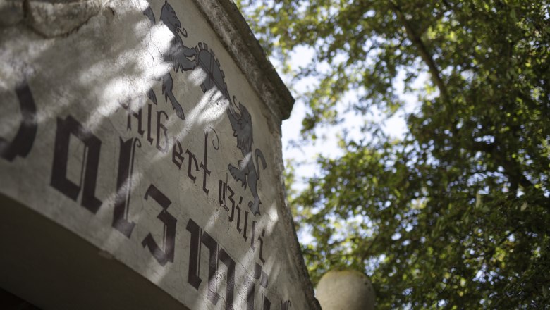 Close-up of an old building with lettering and coat of arms, surrounded by trees.