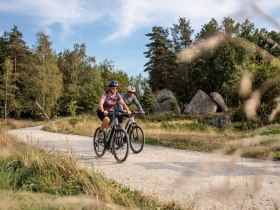 Zwei Radfahrer auf einem Schotterweg im Naturpark Blockheide, umgeben von B&auml;umen und Felsen.